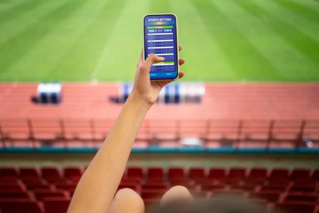 Mão segurando um celular com um aplicativo de apostas, com o campo ao fundo